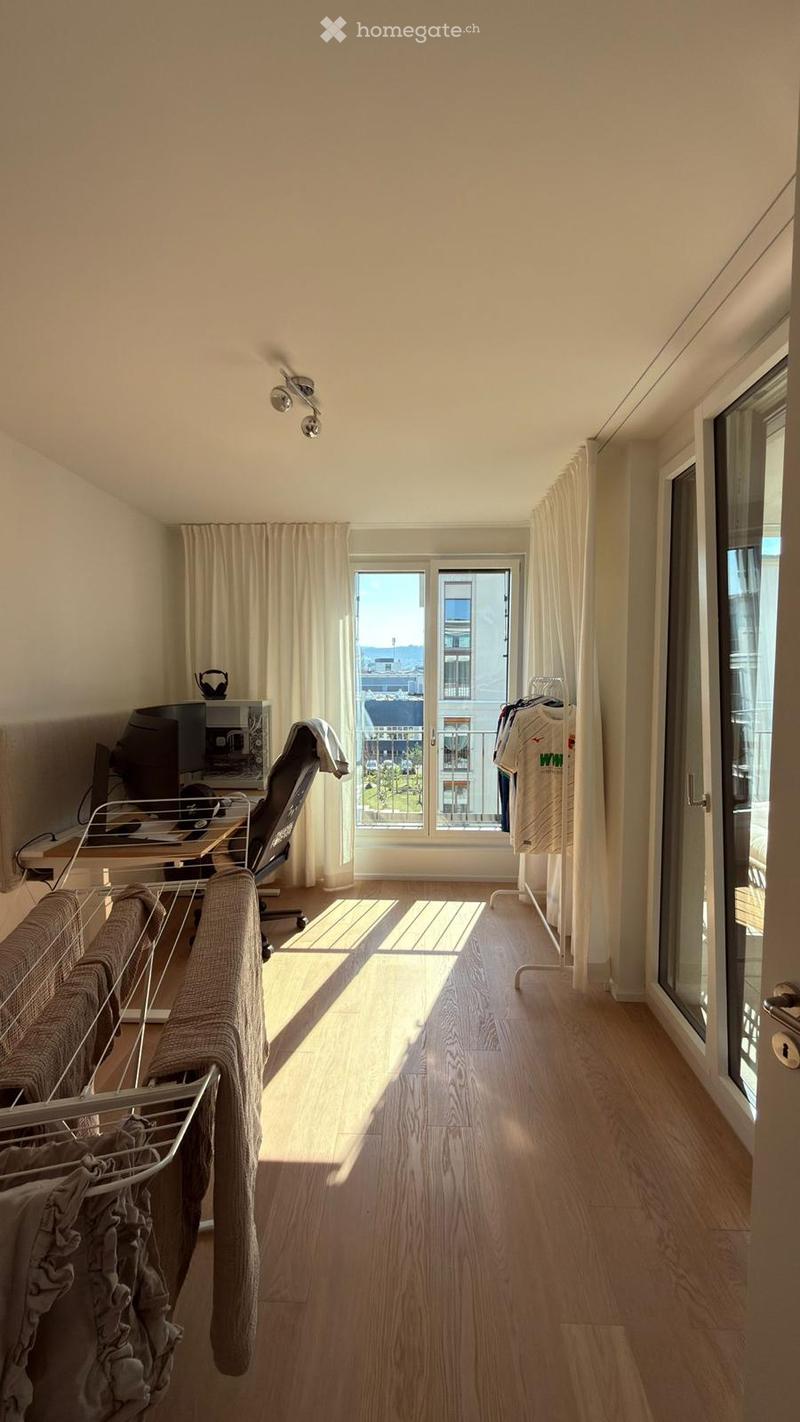 A well-lit room with wooden flooring, large windows, a sofa, a chair, a table with a laptop, and a clothing rack with folded clothes.