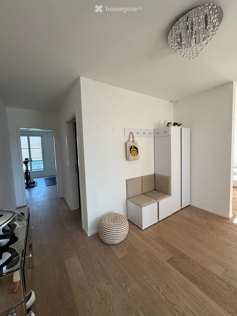 Modern hallway with light wooden flooring, white walls, built-in bench seating, small round ottoman, coat rack with bag, and a decorative chandelier.