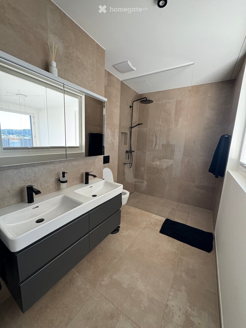 Modern bathroom with beige tiles, double sink, black faucet, large mirror, glass-enclosed shower, and a towel on the floor.