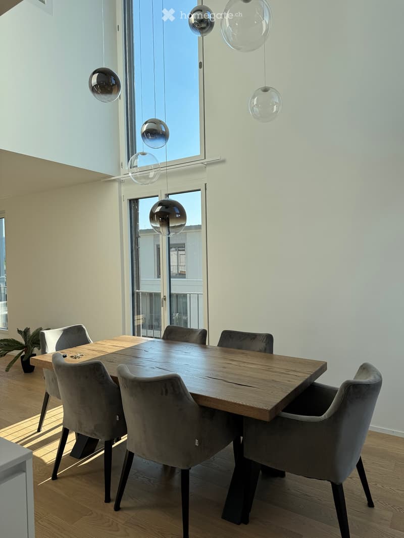 Modern dining area with a wooden table, six grey upholstered chairs, pendant lights, and a small potted plant near the window.