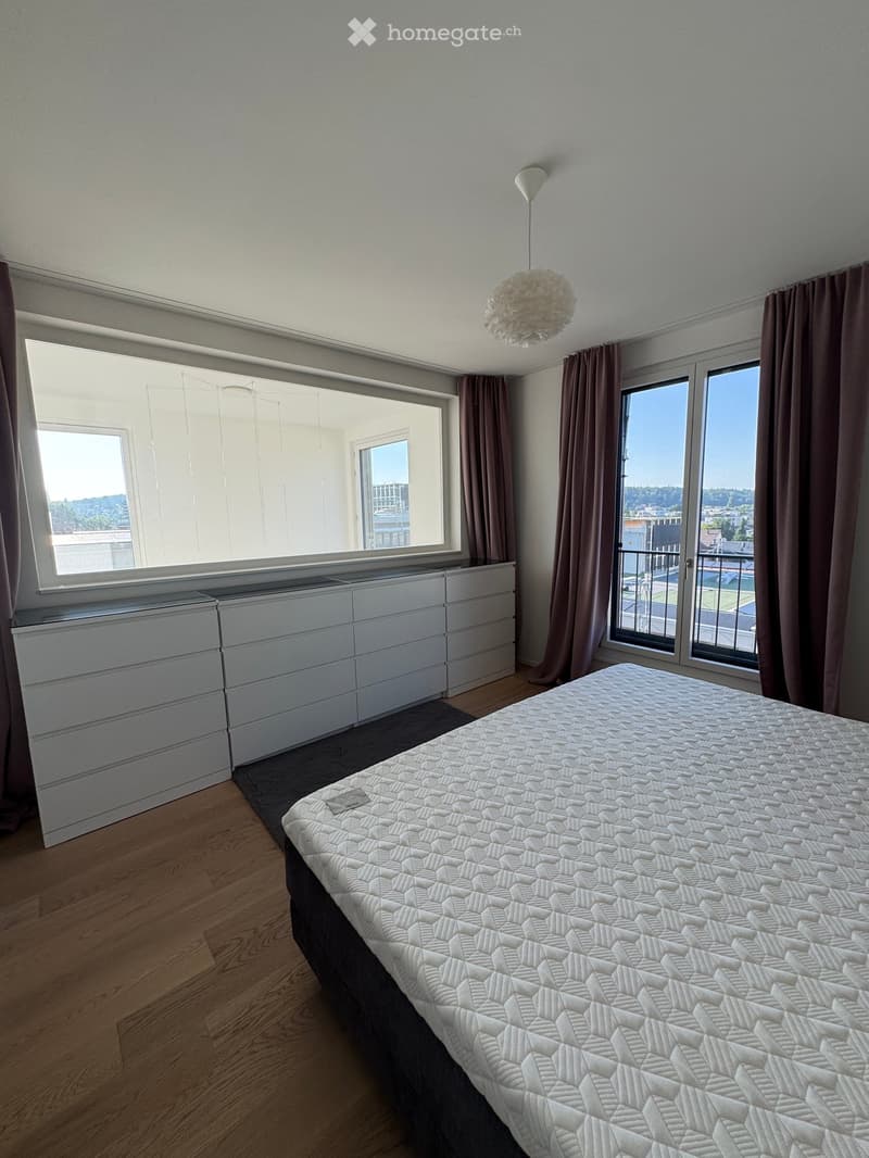 Modern bedroom with a large bed, white dresser, large windows with curtains, and a pendant light fixture.