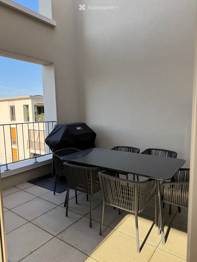 Balcony with a black table, four wicker chairs, and a covered grill, overlooking a cityscape.