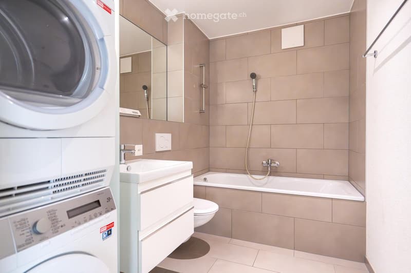 Modern bathroom with beige tiles, a white sink, bathtub, toilet, and a washing machine to the left.