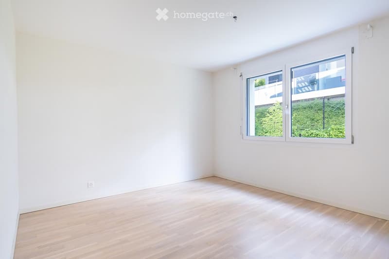 Empty room with white walls and light wooden flooring, featuring a large window with a view of greenery outside.