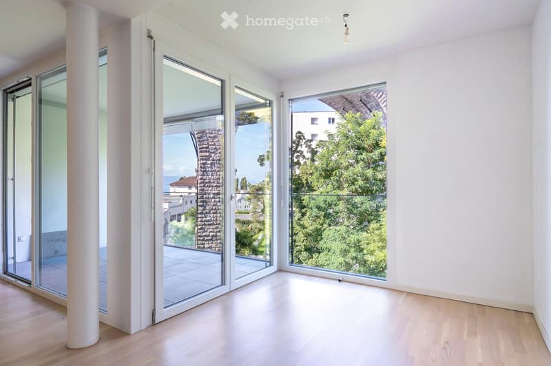 Bright, empty room with large glass doors and windows offering a scenic view of greenery and buildings. Light wooden flooring and white walls.