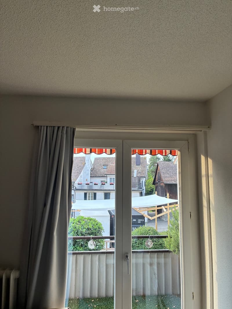 Balcony with white frame, grey curtains, and a view of neighboring houses and a small garden area with a wooden pergola and umbrella.