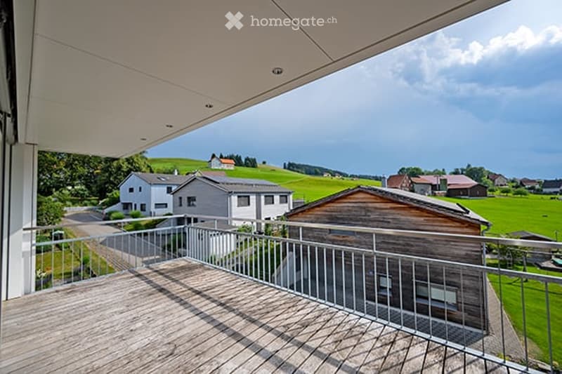 Balcony with wooden decking, metal railing, overlooking a rural landscape with houses and green fields.