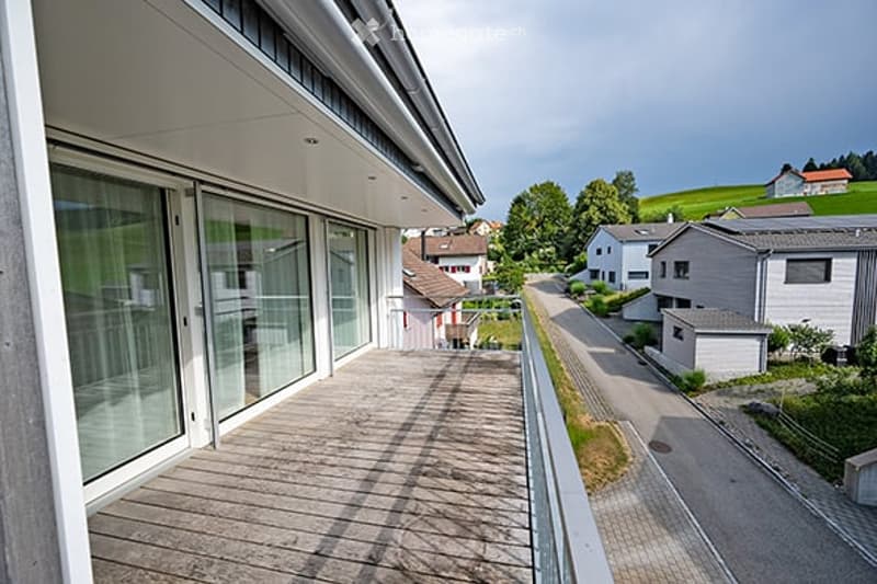Balcony with wooden decking, large glass sliding doors, overlooking a residential street with houses and greenery.