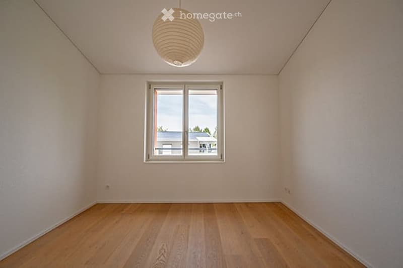 Empty room with light-colored walls, wooden flooring, a single window, and a hanging light fixture.