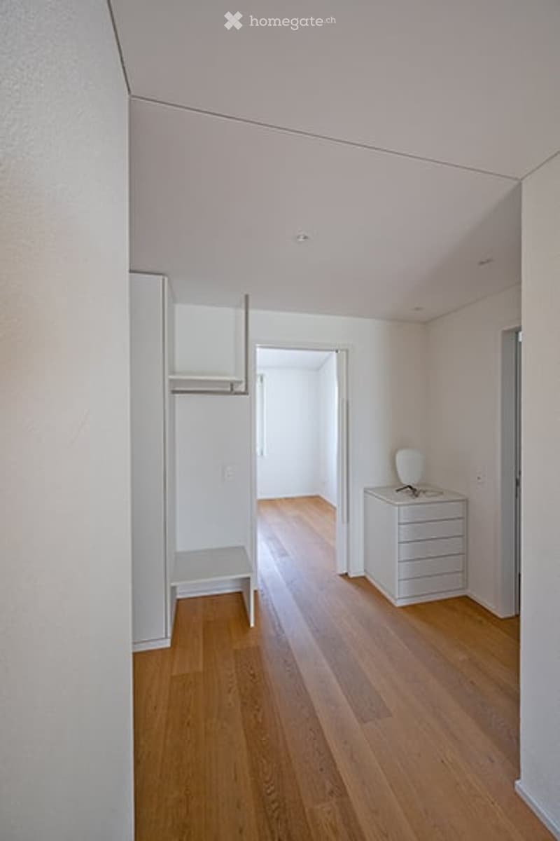 A small, minimalist room with light wooden flooring, white walls, and a single chair and drawer unit.