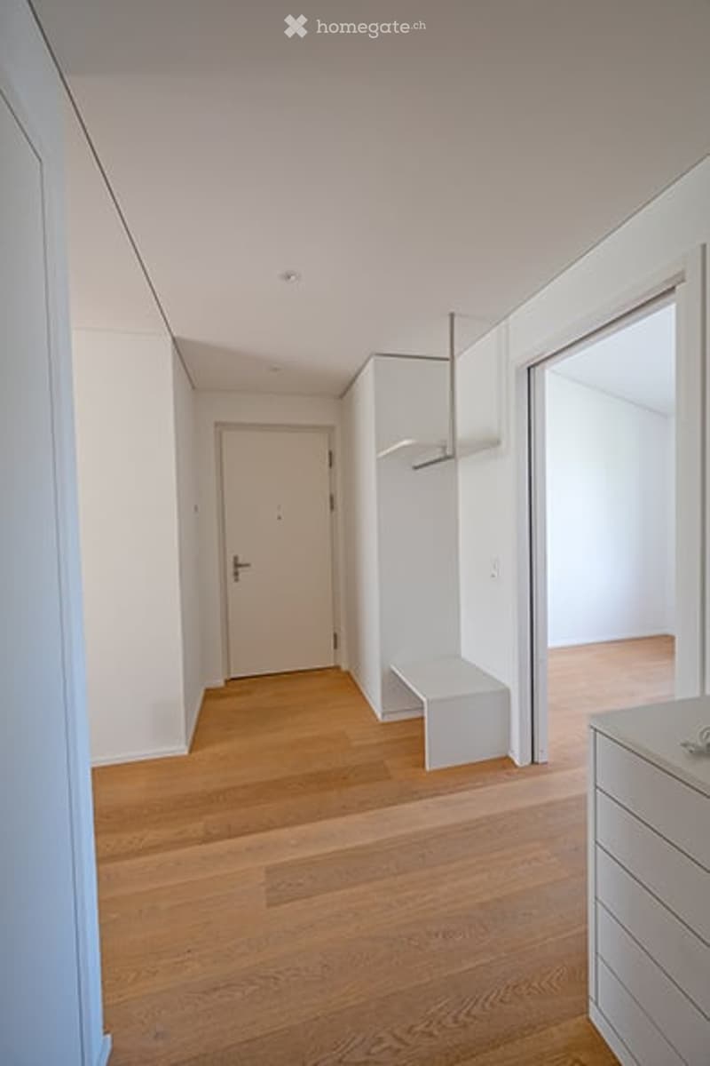 Empty hallway with light wooden flooring, white walls, and a closed white door at the end. A small white bench is placed against the wall near the door.