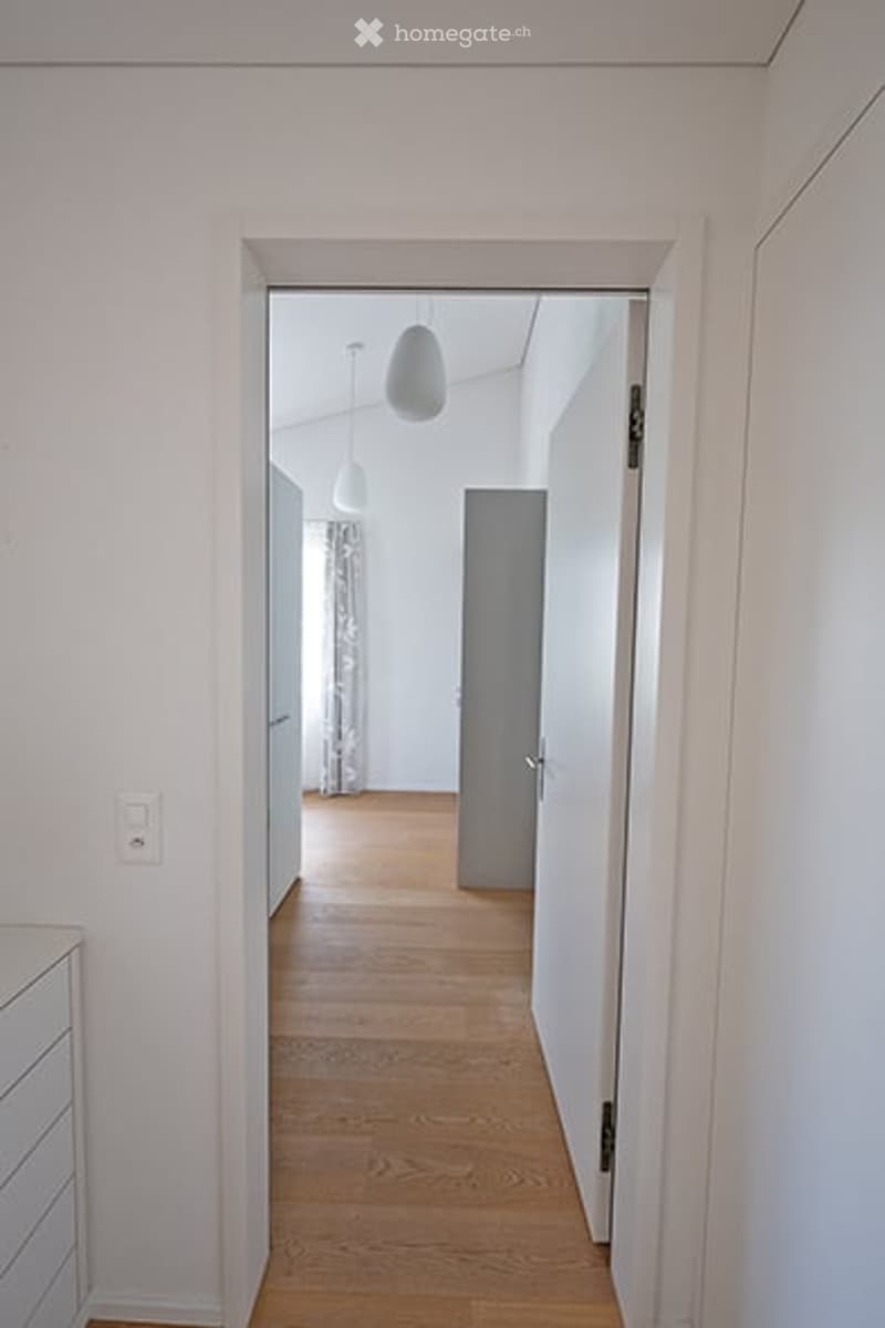 A bright, minimalist hallway with light wooden flooring, white walls, and pendant lights. The hallway leads to an open area with a visible curtain and a closed door.