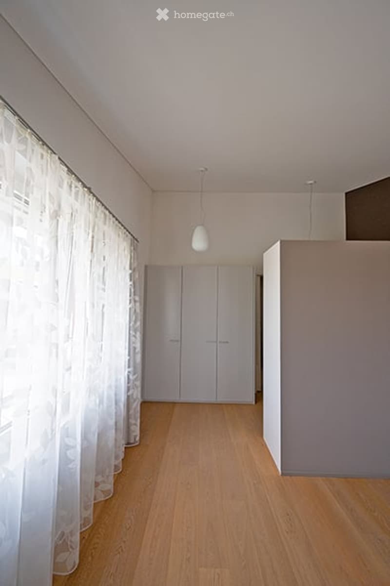 Empty room with light wooden flooring, white walls, built-in closet, and white curtains on the window. No furniture or personal items present.