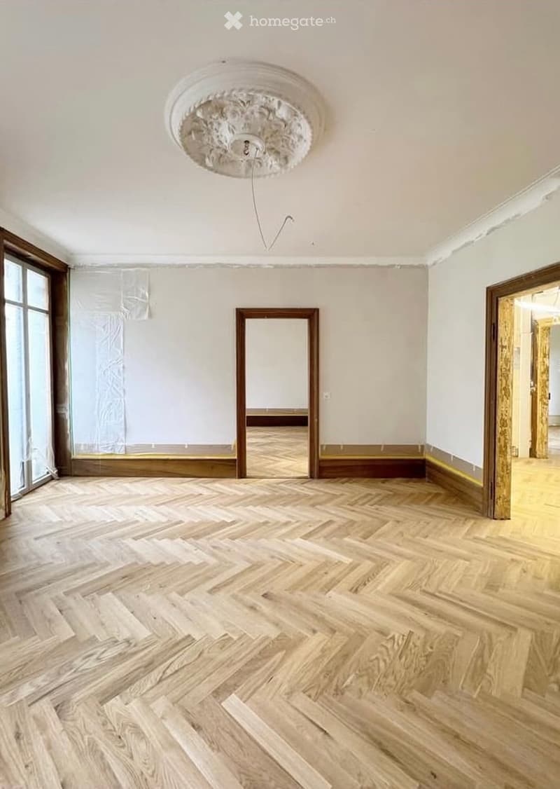 Empty room with wooden herringbone flooring, large windows, and a decorative ceiling light fixture.