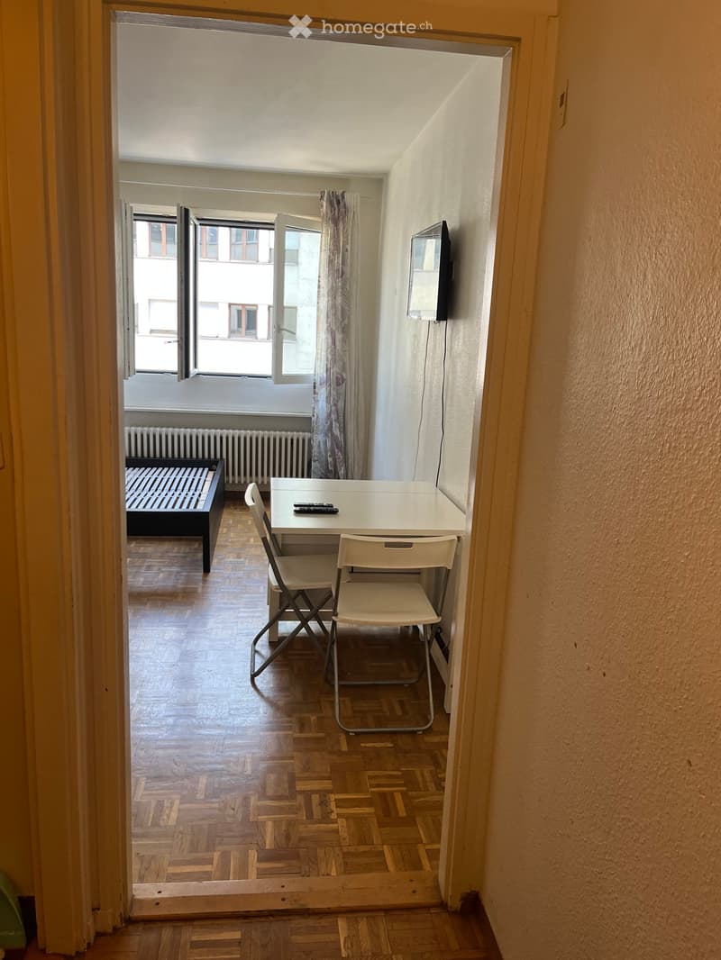 Small furnished room with wooden flooring, a bed, a table with two chairs, and a television mounted on the wall. The room has large windows with open shutters, allowing natural light to enter.