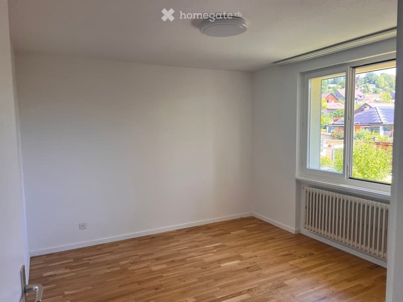 Empty room with light-colored walls, wooden flooring, a window with a view of the neighborhood, and a radiator below the window.