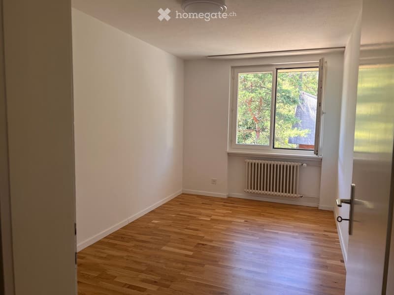 Empty room with wooden flooring, white walls, a window with a view of trees, and a radiator below the window.