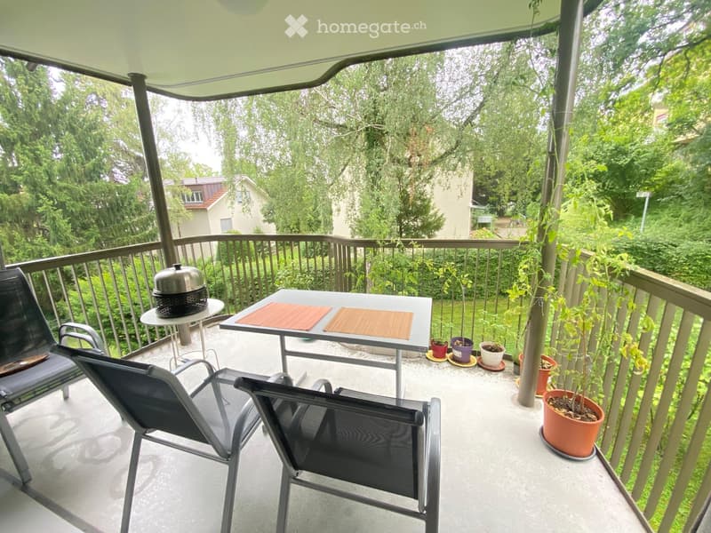 Balcony with a table, chairs, and potted plants, surrounded by greenery and a covered roof.