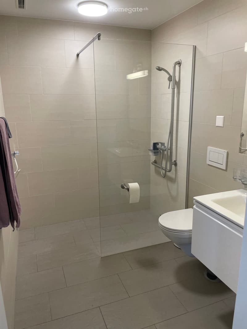 Modern bathroom with beige tiles, a glass-enclosed shower, a toilet, a sink, and a towel hanging on the wall.