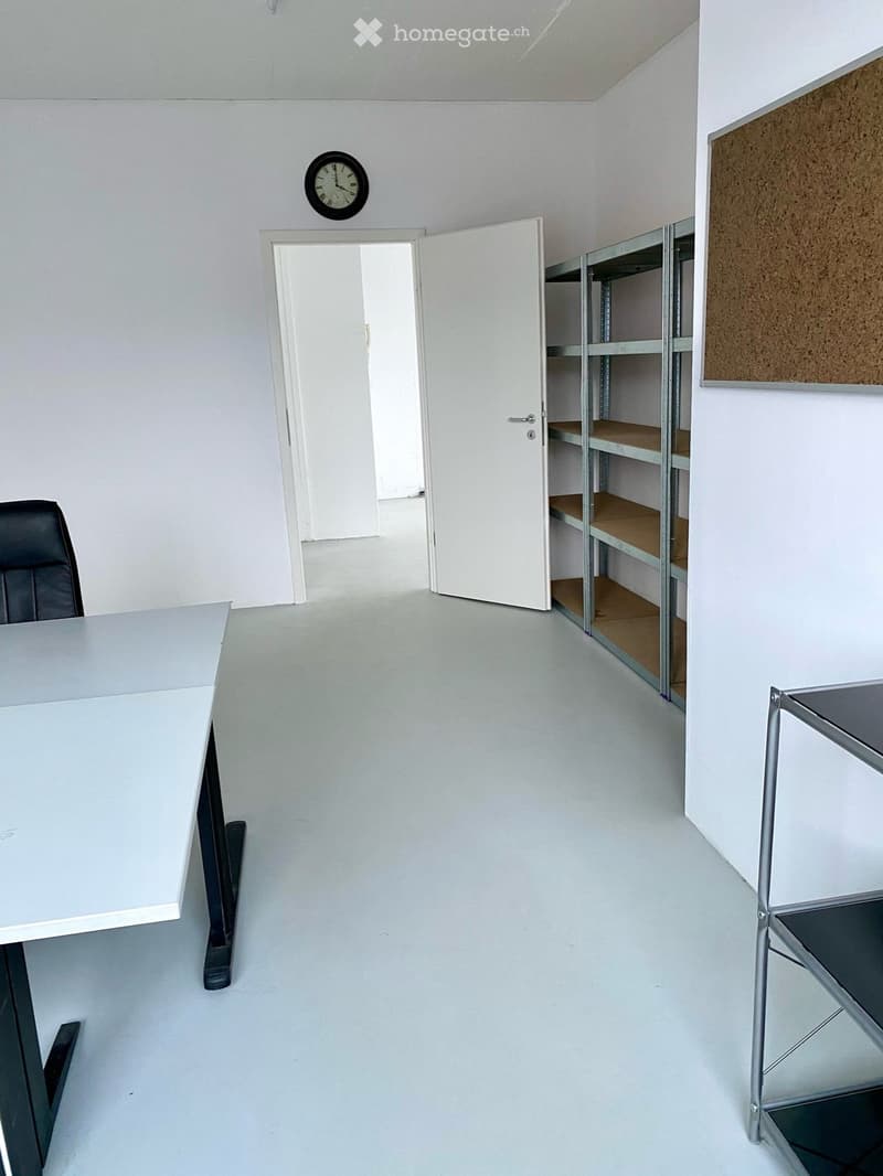 A clean, minimalist office space with a white table, black chair, metal shelving unit, and a wall-mounted clock.