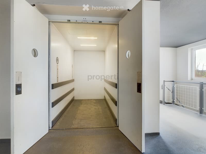 Empty hallway with white walls, gray flooring, and a large open doorway leading to another area. The hallway features a small window on the right side and a metal railing leading to a staircase or balcony area.
