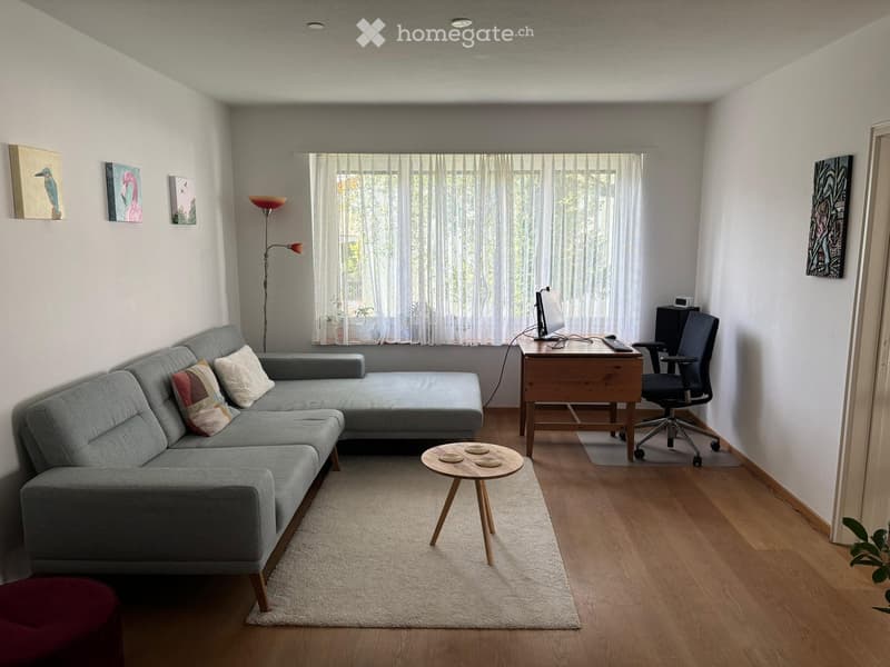 Modern living room with a grey sectional sofa, wooden coffee table, desk with computer, and floor lamp. The room features large windows with sheer curtains and a small round wooden stool.