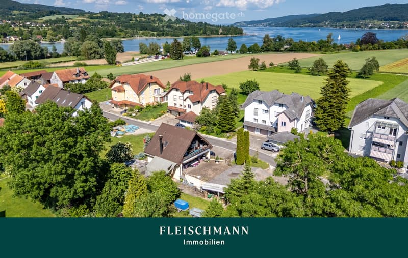 Aerial view of a residential area with multiple houses, surrounded by green fields, trees, and a lake in the background.