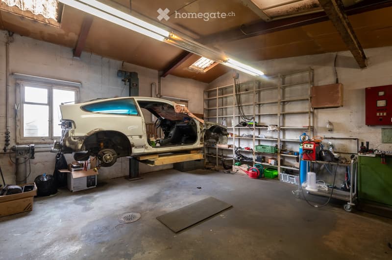 A spacious garage with a partially disassembled white car on a lift, surrounded by shelves filled with tools and equipment, and a workbench with various items.