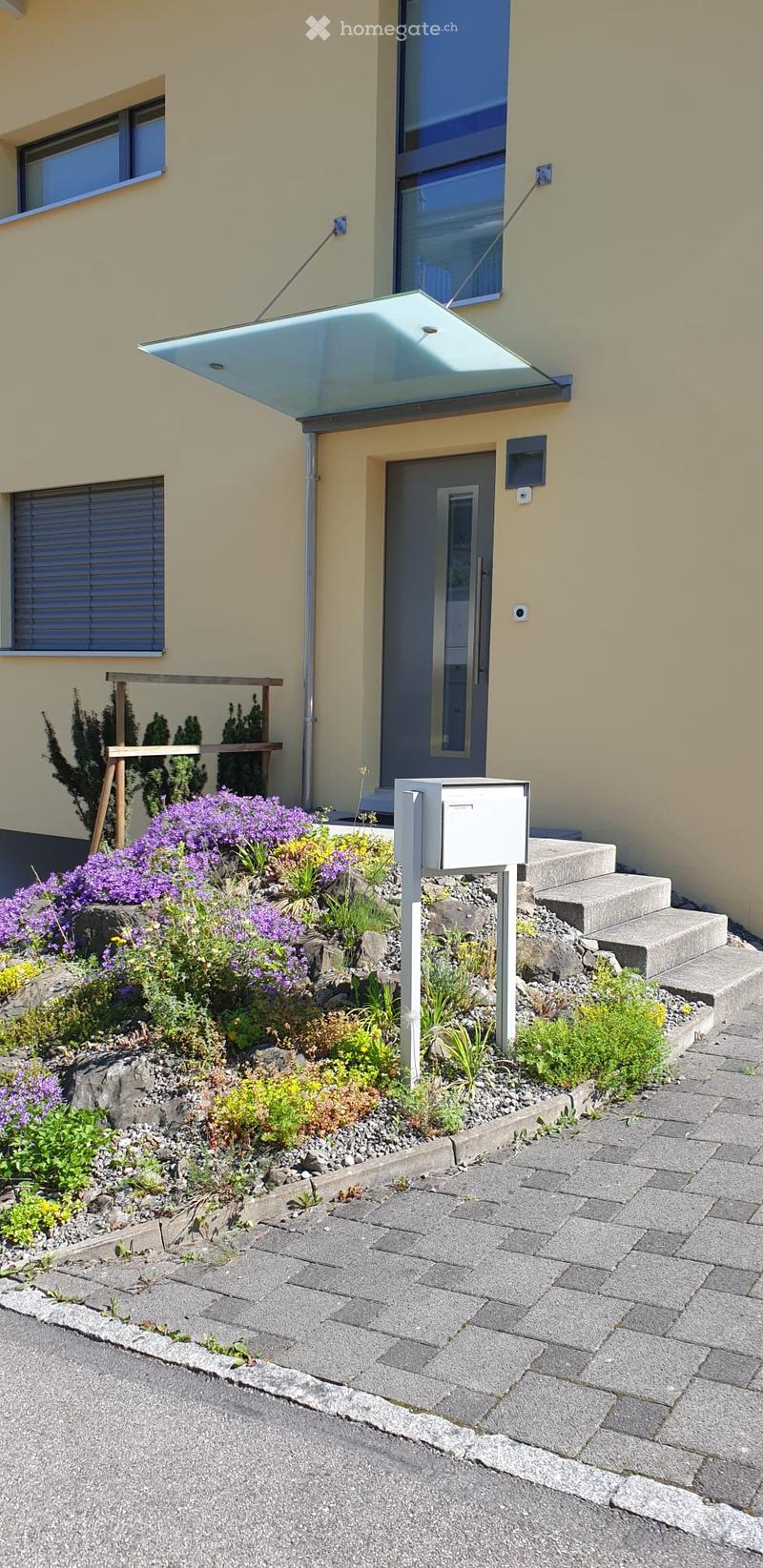 Front garden with colorful flowers, shrubs, and a mailbox, leading to a modern house entrance with a glass canopy and steps.