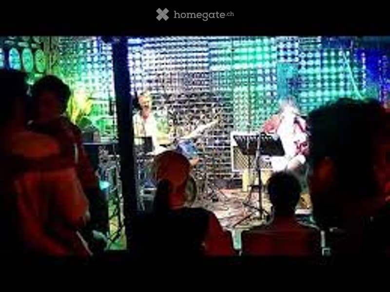 A live music performance in a studio setting with a band playing instruments and a small audience watching.