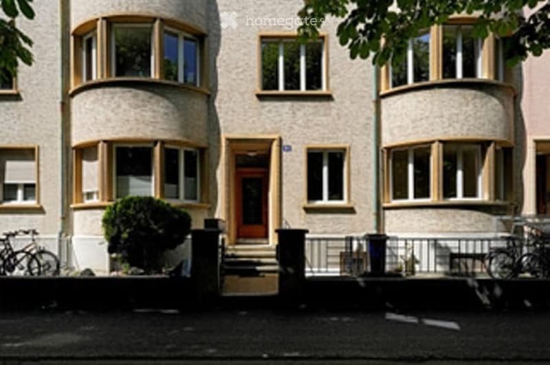 Raised ground floor, two-story building, beige exterior with yellow window frames, multiple arched windows, two bicycles parked in front