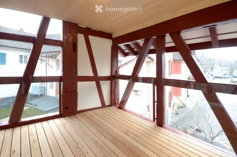 Wooden deck with glass railings, exposed wooden beams, and a view of the street and neighboring buildings.