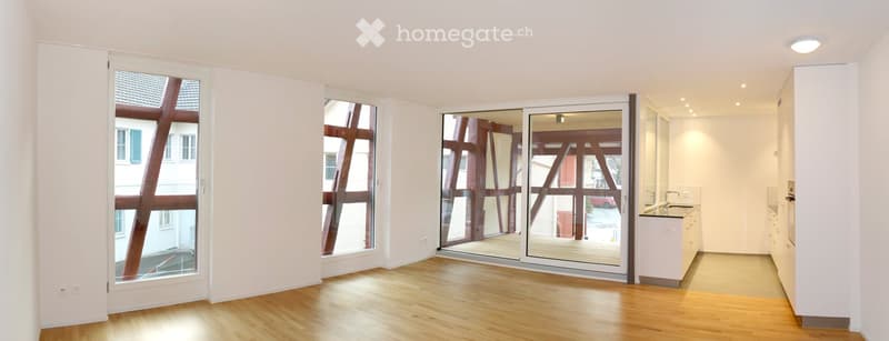 Empty room with wooden flooring, large windows, and a sliding glass door leading to a balcony area. The kitchen area is visible with white cabinets and a sink.