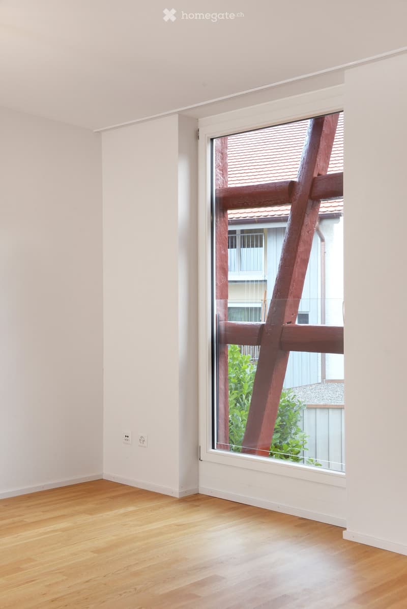 Empty room with light-colored wooden flooring and white walls, featuring a large window with a view of a wooden structure and greenery outside.