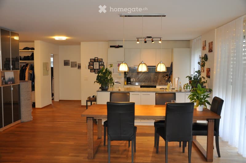 Dining room with wooden table, black chairs, wooden flooring, open to kitchen, ceiling lamps, plants, and framed pictures on the walls.