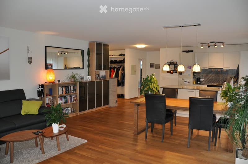 Wooden floors, wooden dining table, 4 black chairs, wooden side tables, grey couch with green pillow, lamps, plants, open kitchen with countertops