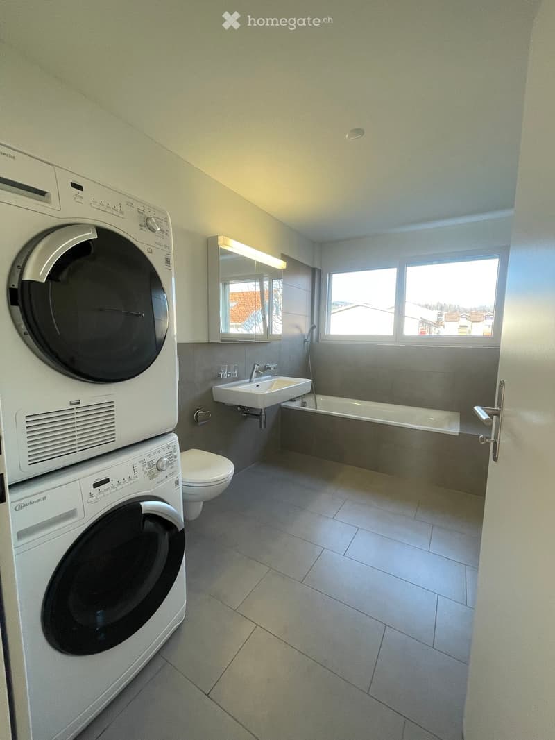 A bathroom with a white bathtub, sink, toilet, washing machines, and a large window