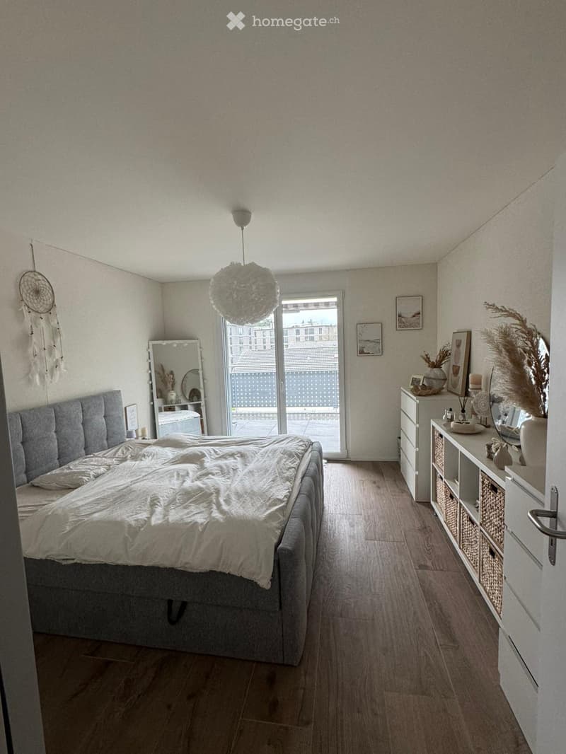 large bed with grey headboard, wooden floor, white walls, balcony with railing, full-length mirror, decorative items, and cabinets