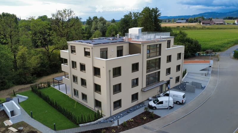 Modern multi-story building with solar panels on the roof, surrounded by a landscaped garden and parking area.