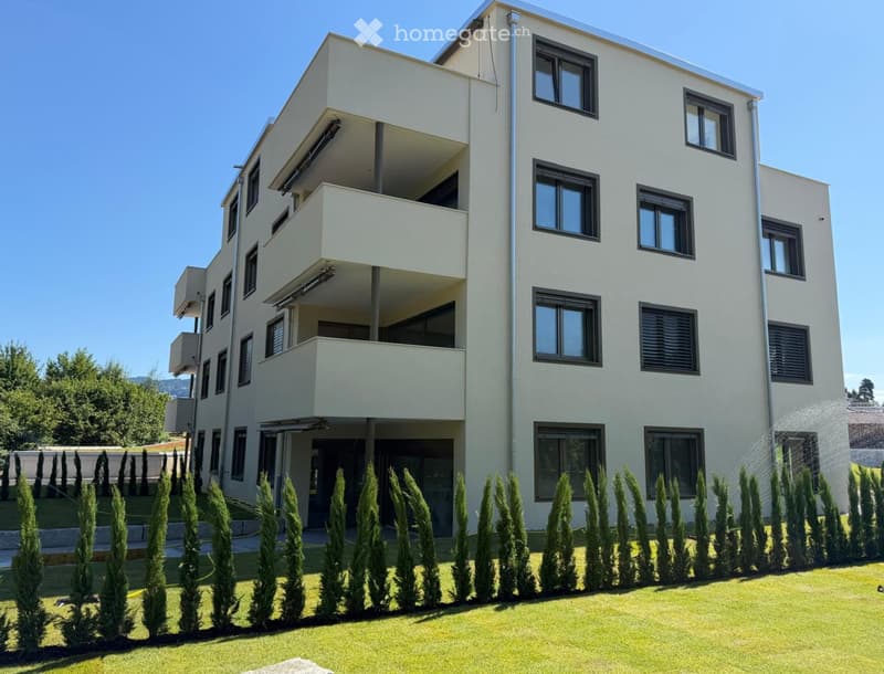 Modern three-story building with white facade, multiple windows, balconies, and surrounding greenery.