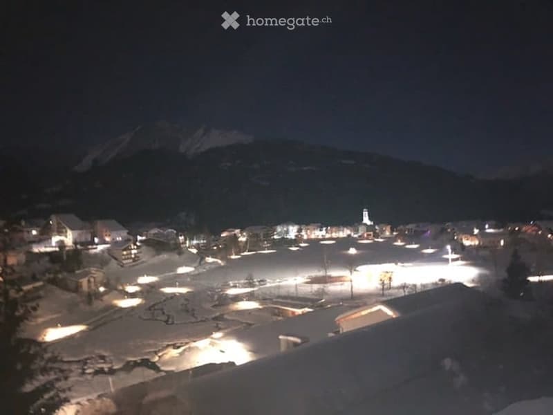 Nighttime view of a snow-covered village with illuminated buildings and a distant mountain range.