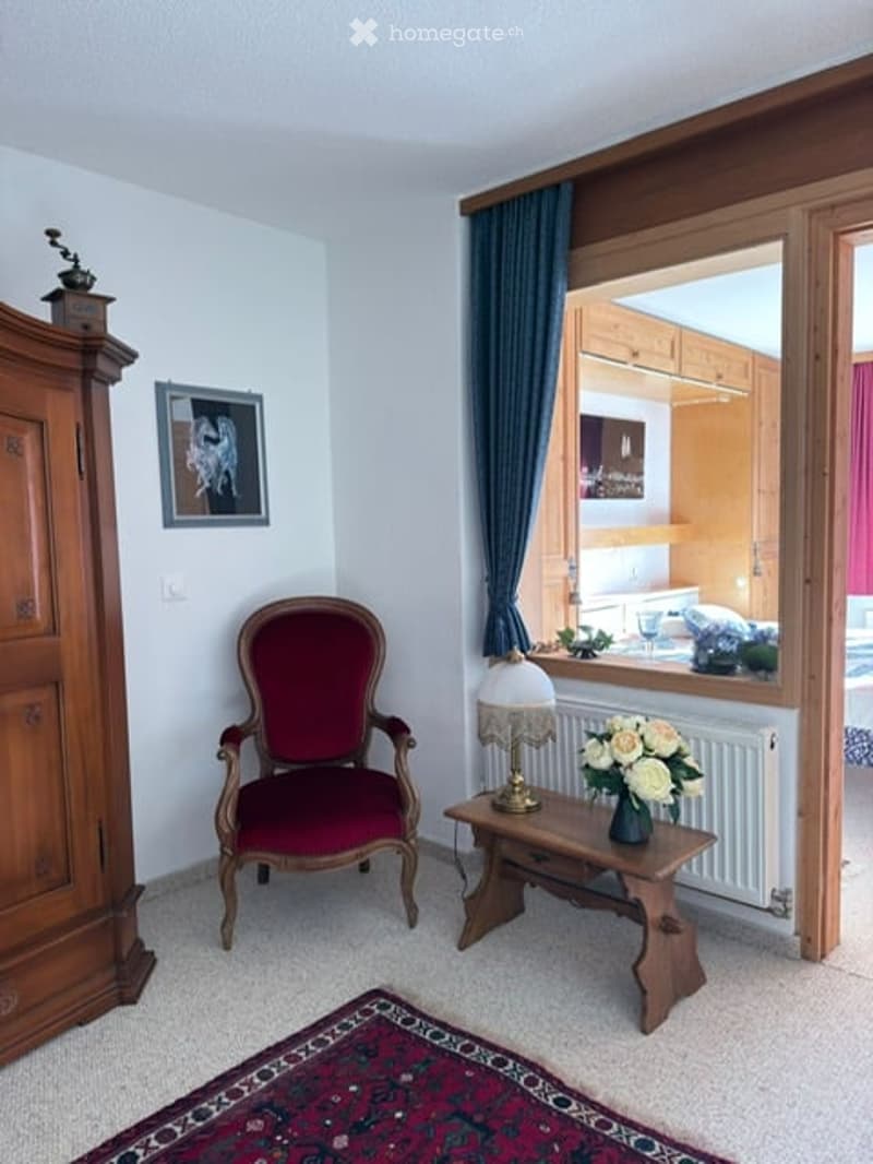 A cozy, furnished room with a red upholstered chair, a small wooden table with a lamp and flower vase, a wooden wardrobe, and a decorative picture frame on the wall. The room has large windows with blue curtains and a view of another room.