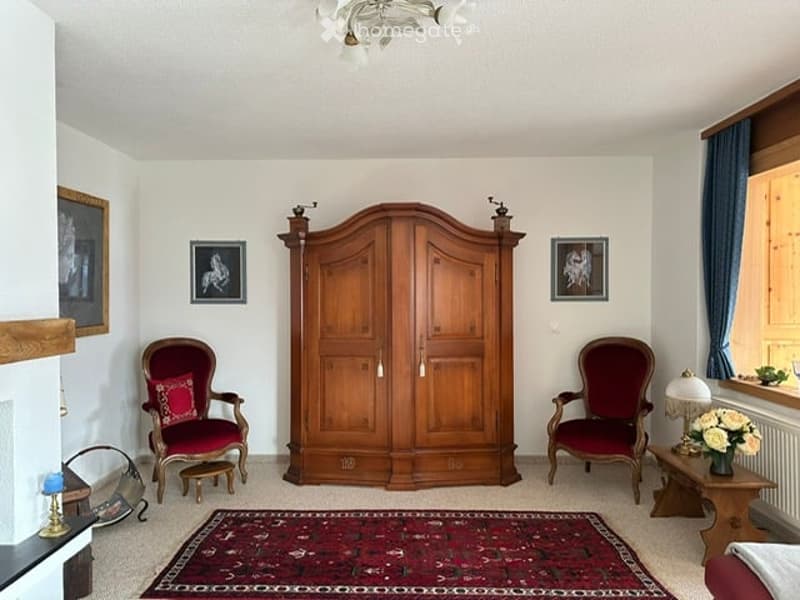 Elegant living room with two red armchairs, a large wooden wardrobe, a small table with a vase of flowers, and several picture frames on the walls.