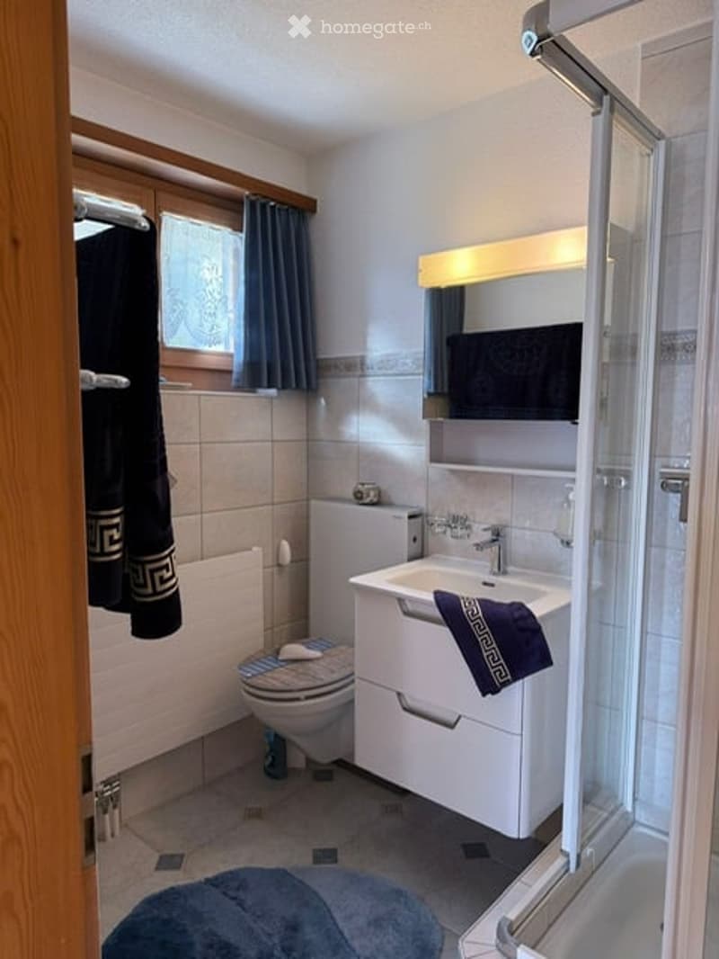 Modern bathroom with white sink, mirror, toilet, and shower. Blue towels and a dark curtain add a touch of color.