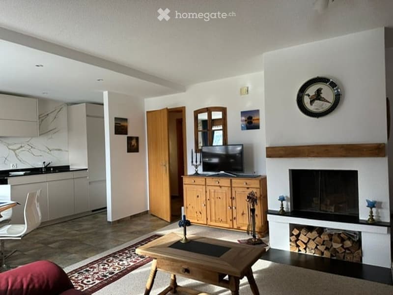 Bright living room with a fireplace, wooden coffee table, sofa, TV on a wooden cabinet, and a mirror above the fireplace. The room features a mix of modern and rustic elements, including a patterned rug and a dining area with white chairs.