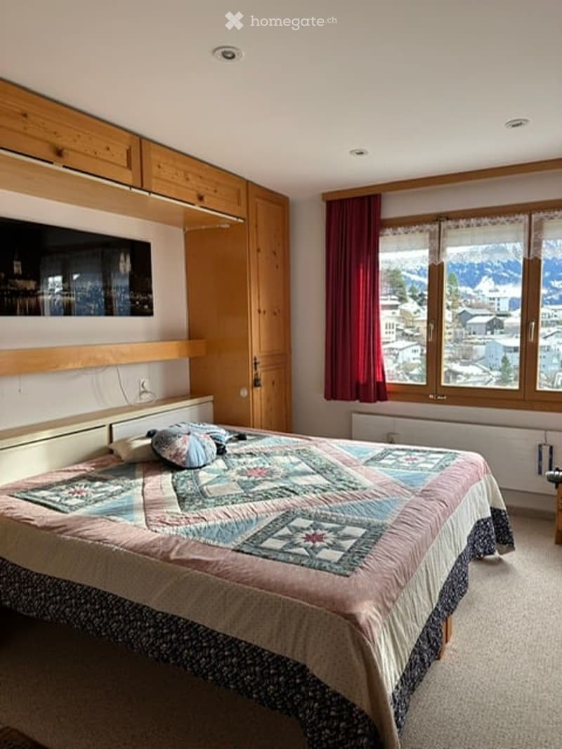 Cozy bedroom with a large window offering a scenic mountain view, featuring a neatly made bed with pillows and a colorful quilt, a wooden wardrobe, and a mirror on the wall.