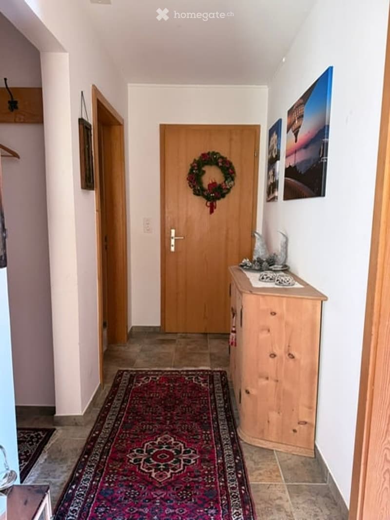Hallway with wooden doors, a decorative wreath on one door, a small wooden cabinet with items on top, and a patterned rug on the floor.