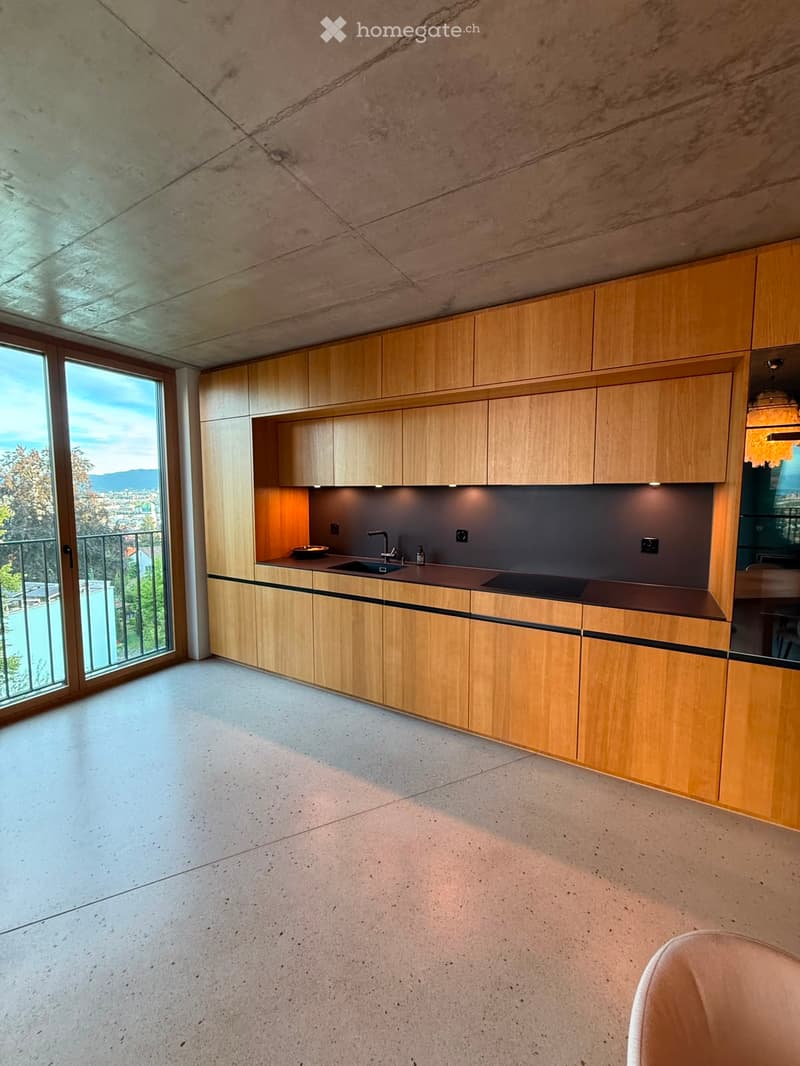 Modern kitchen with wooden cabinets, concrete floor, and large windows offering a scenic view.