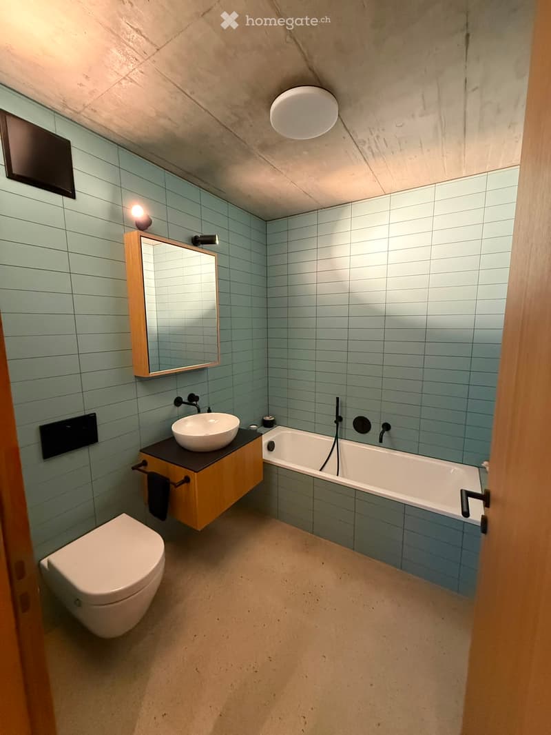 Modern bathroom with light blue tiled walls, a freestanding bathtub, a wall-mounted sink with a wooden vanity, a toilet, and a large mirror with integrated lighting.
