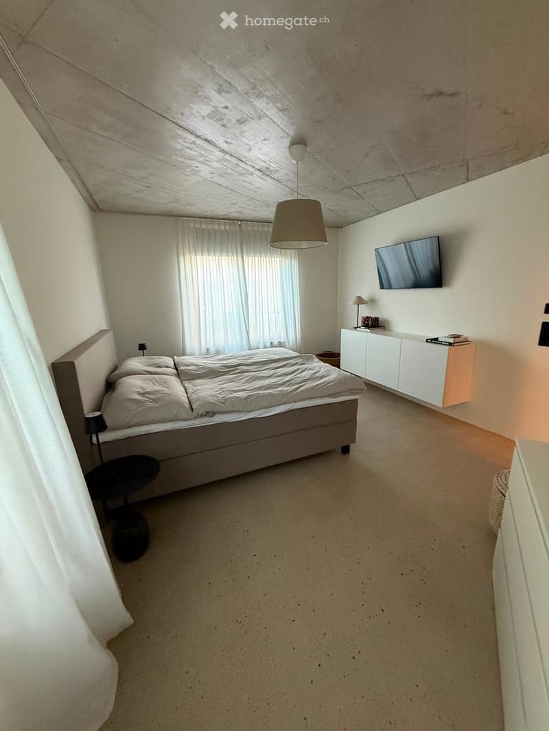 Modern bedroom with a large bed, nightstand, table lamp, and television mounted on the wall. The room features large windows with curtains and a sleek, minimalist design.